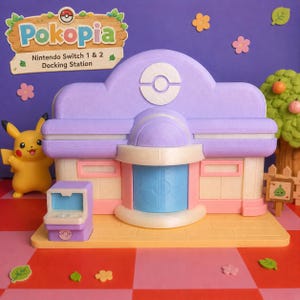May include: A multi-colored Nintendo Switch 1 & 2 docking station, designed as a building with a light purple roof and a blue door. The station has a yellow base and is decorated with flowers and a Pikachu figure. The text "Pokopia" is visible.