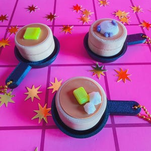 May include: Three miniature pancake charms with a pat of green butter on top, each in a tiny black frying pan. The pancakes are tan with a brown top, and the pans have a blue handle. Gold star confetti is scattered on a pink surface.