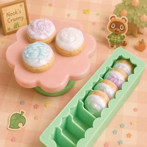 Pode incluir: Miniaturas de sobremesas decoradas, tr&ecirc;s em um suporte rosa em forma de flor e cinco em um recipiente verde. As sobremesas t&ecirc;m cobertura branca, azul e rosa. Uma placa diz "Nook's Cranny".