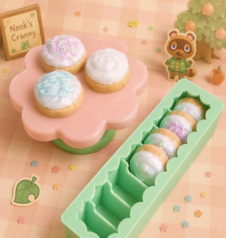 Pode incluir: Miniaturas de sobremesas decoradas, tr&ecirc;s em um suporte rosa em forma de flor e cinco em um recipiente verde. As sobremesas t&ecirc;m cobertura branca, azul e rosa. Uma placa diz "Nook's Cranny".