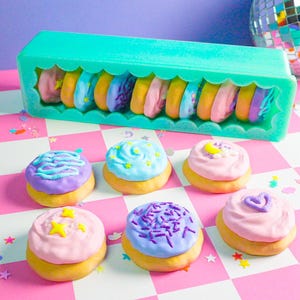 Pode incluir: Uma cole&ccedil;&atilde;o de mini-donuts coloridos e decorados. Alguns est&atilde;o em um recipiente turquesa, outros est&atilde;o dispostos em uma superf&iacute;cie xadrez rosa e branca. Os donuts t&ecirc;m v&aacute;rias coberturas e granulados em tons pastel.