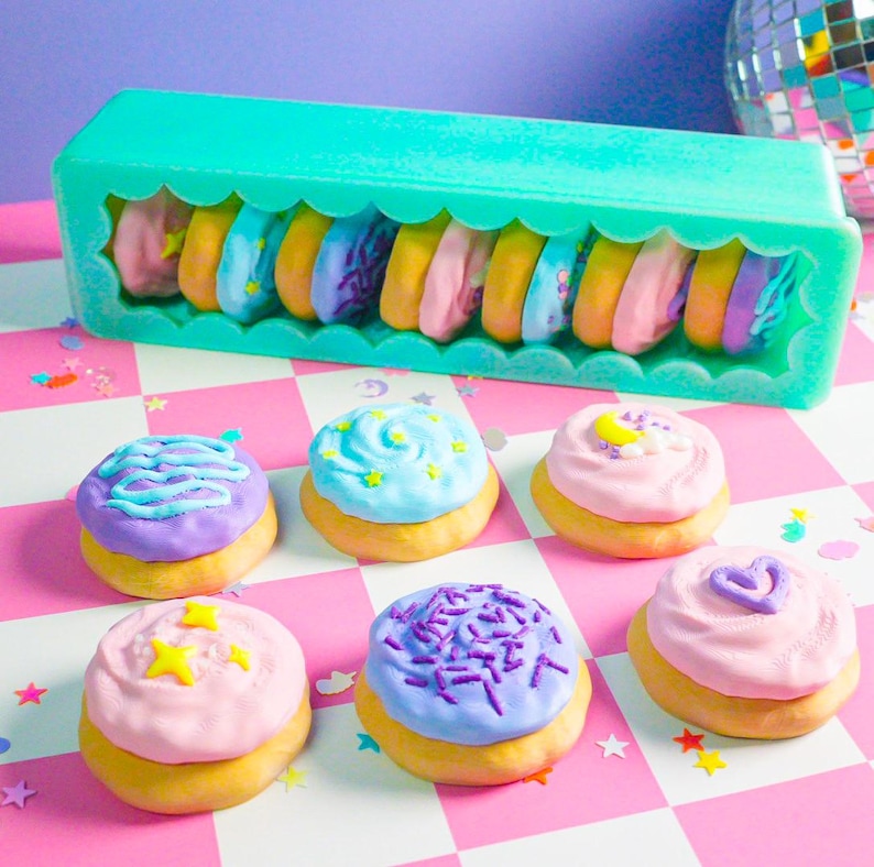 Pode incluir: Uma cole&ccedil;&atilde;o de mini-donuts coloridos e decorados. Alguns est&atilde;o em um recipiente turquesa, outros est&atilde;o dispostos em uma superf&iacute;cie xadrez rosa e branca. Os donuts t&ecirc;m v&aacute;rias coberturas e granulados em tons pastel.