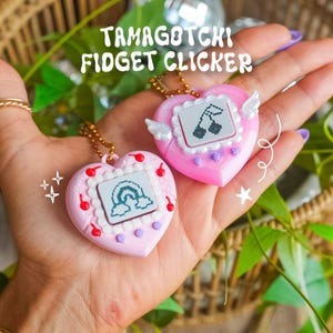 May include: Two heart-shaped Tamagotchi fidget clickers with gold chains. One is light pink with a rainbow screen graphic and red cherry accents. The other is bright pink with a cherry screen graphic and white wings on the sides.