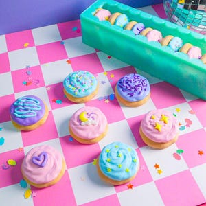 Pode incluir: Mini donuts coloridos com cobertura pastel e decora&ccedil;&otilde;es de granulados, estrelas e cora&ccedil;&otilde;es. Os donuts est&atilde;o dispostos sobre uma superf&iacute;cie xadrez rosa e branca. Um recipiente retangular verde cont&eacute;m mais donuts no fundo.