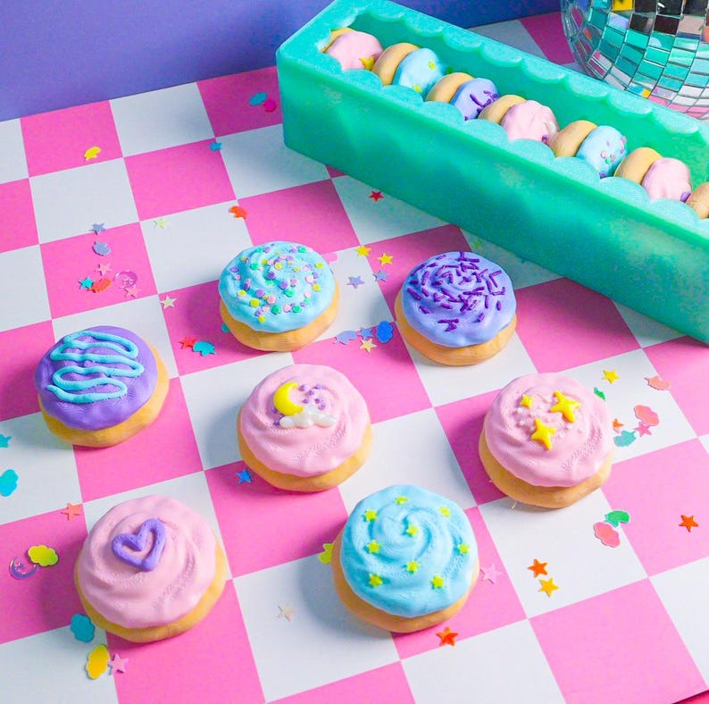 Pode incluir: Mini donuts coloridos com cobertura pastel e decora&ccedil;&otilde;es de granulados, estrelas e cora&ccedil;&otilde;es. Os donuts est&atilde;o dispostos sobre uma superf&iacute;cie xadrez rosa e branca. Um recipiente retangular verde cont&eacute;m mais donuts no fundo.