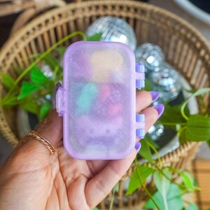 Puede incluir: Un peque&ntilde;o contenedor de almacenamiento rectangular, transl&uacute;cido y de color morado claro con un acabado brillante y una tapa con bisagras. La caja cuenta con un mecanismo de cierre seguro en el lateral y contiene varios objetos peque&ntilde;os y coloridos.