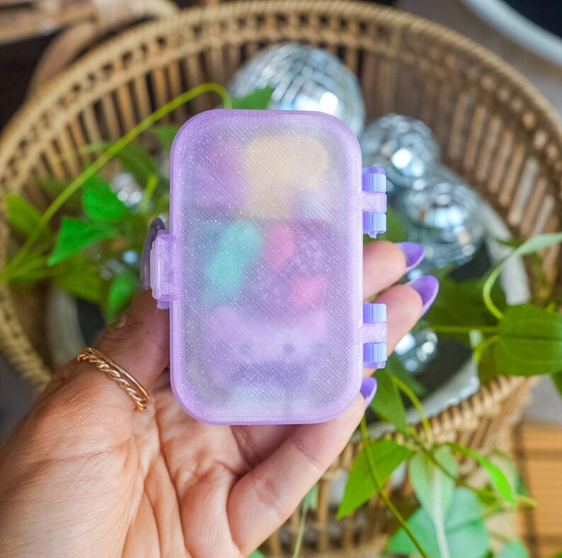 Puede incluir: Un peque&ntilde;o contenedor de almacenamiento rectangular, transl&uacute;cido y de color morado claro con un acabado brillante y una tapa con bisagras. La caja cuenta con un mecanismo de cierre seguro en el lateral y contiene varios objetos peque&ntilde;os y coloridos.