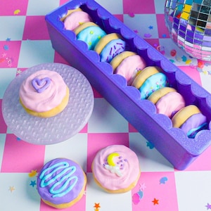 Pode incluir: Uma cole&ccedil;&atilde;o de biscoitos coloridos com cobertura rosa, azul e roxa. Os biscoitos s&atilde;o dispostos em um recipiente roxo com bordas onduladas e em um prato texturizado. O fundo apresenta um padr&atilde;o xadrez rosa e branco.