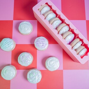 Pode incluir: Uma cole&ccedil;&atilde;o de biscoitos com cobertura e macarons em um recipiente rosa, dispostos em uma superf&iacute;cie xadrez vermelha e rosa. Os biscoitos t&ecirc;m cobertura em tons pastel, e os macarons t&ecirc;m um esquema de cores marrom claro e branco.