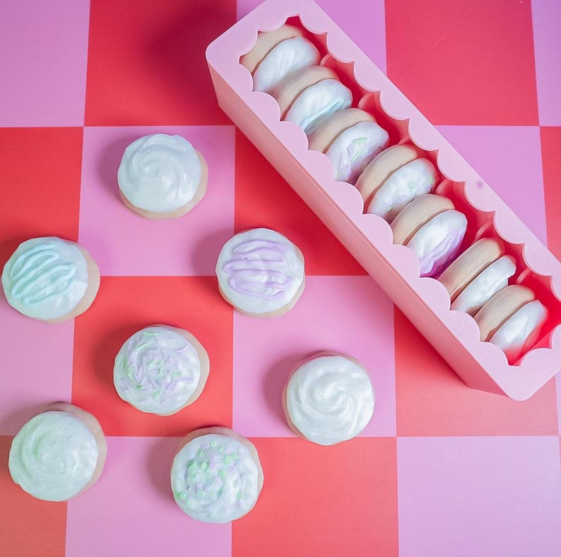 Pode incluir: Uma cole&ccedil;&atilde;o de biscoitos com cobertura e macarons em um recipiente rosa, dispostos em uma superf&iacute;cie xadrez vermelha e rosa. Os biscoitos t&ecirc;m cobertura em tons pastel, e os macarons t&ecirc;m um esquema de cores marrom claro e branco.
