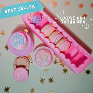 May include: A pink cookie-shaped pill organizer with the text "COOKIE PILL ORGANIZER" and "BEST SELLER". The organizer holds several cookie-shaped pills with pink, blue, and yellow frosting. Additional cookie-shaped containers are also visible.