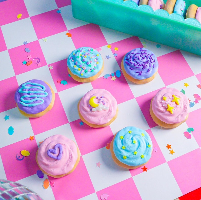 Pode incluir: Uma cole&ccedil;&atilde;o de donuts coloridos com cobertura e v&aacute;rias coberturas, incluindo granulado, redemoinhos e formas de estrela. Os donuts est&atilde;o dispostos sobre uma superf&iacute;cie quadriculada rosa e branca, com confetes espalhados.