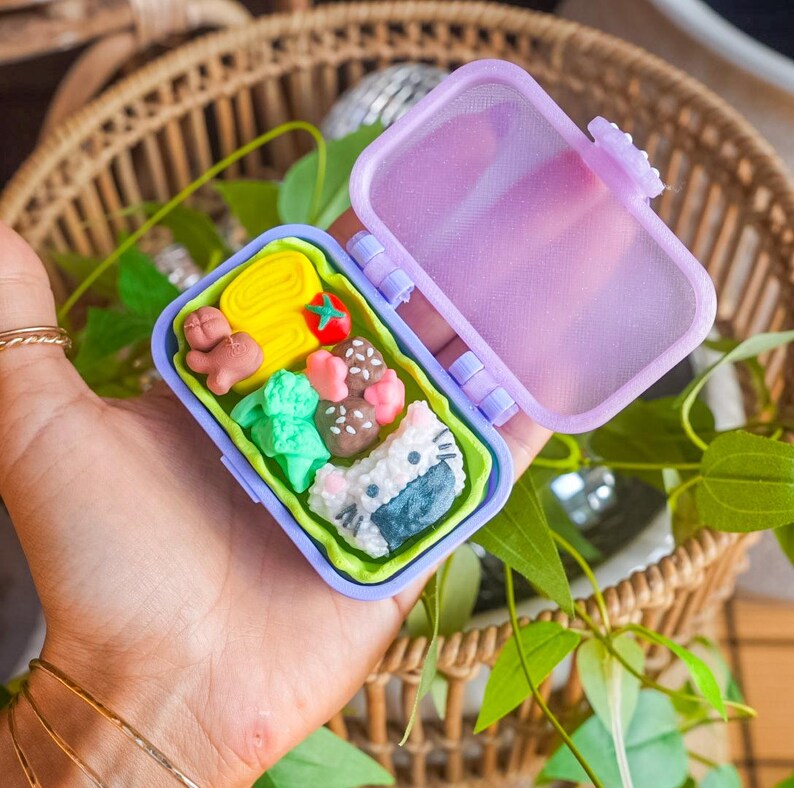 Puede incluir: Una peque&ntilde;a caja bento rectangular de pl&aacute;stico morado claro con la tapa abierta. En su interior hay gomas de borrar en miniatura con forma de comida, incluyendo una tortilla amarilla, un tomate rojo, una salchicha marr&oacute;n y br&oacute;coli verde.