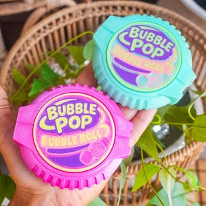 May include: Two round plastic bubble gum dispensers, one in bright pink and one in light mint green. Each features a circular label with the text BUBBLE POP BUBBLE ROLL in purple and yellow, along with a graphic of three small bubble gum spheres.