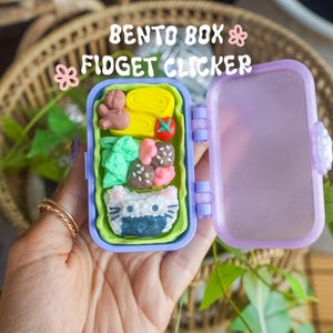 Puede incluir: Una caja bento fidget clicker de pl&aacute;stico morado con la tapa abierta. Dentro hay alimentos en miniatura que incluyen una tortilla amarilla, un tomate rojo, br&oacute;coli verde, alb&oacute;ndigas marrones y una bola de arroz blanca con forma de gato.