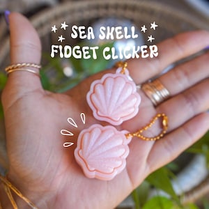 May include: Two light pink, glittery seashell-shaped fidget clickers with gold-toned ball chains. The text SEA SHELL FIDGET CLICKER is printed in white above the items, accompanied by small star and motion line graphics.