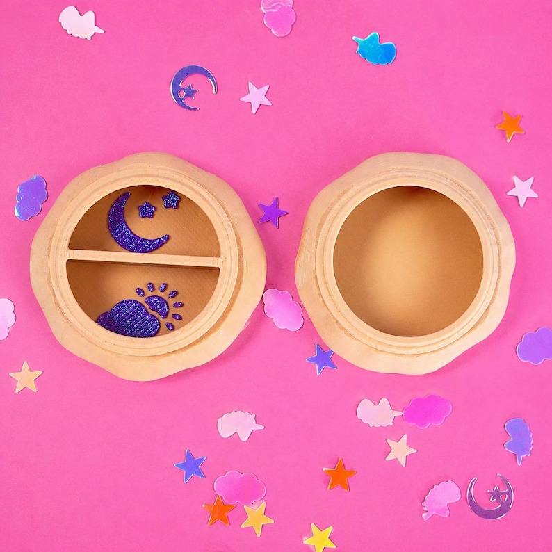 Pode incluir: Duas caixas redondas de madeira com acabamento castanho claro sobre um fundo rosa, uma aberta e outra fechada. A caixa fechada apresenta um desenho de lua crescente e sol em glitter azul. Decora&ccedil;&otilde;es em forma de estrela e nuvem espalhadas.