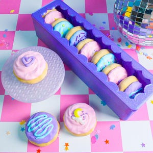 Pode incluir: Uma cole&ccedil;&atilde;o de biscoitos coloridos e com cobertura em um recipiente roxo com bordas onduladas e em um prato transparente. Os biscoitos t&ecirc;m cobertura rosa, azul e roxa com granulados e formas decorativas. O fundo &eacute; um padr&atilde;o xadrez rosa e branco.