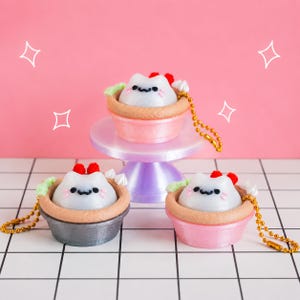 May include: Three cat-shaped charms in tart-like holders, each with a gold ball chain. The charms feature white cat faces with black eyes, pink whiskers, and red and green accents. One charm sits on a small pedestal, while two sit below.