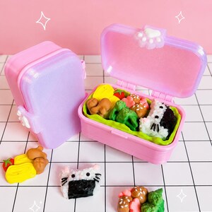 Puede incluir: Dos cajas bento de pl&aacute;stico rosa en miniatura con tapas brillantes y un cierre de huella blanca. Una caja est&aacute; abierta y muestra peque&ntilde;as gomas de borrar con forma de comida, incluyendo br&oacute;coli, bolas de arroz y carne. Hay gomas sueltas cerca.