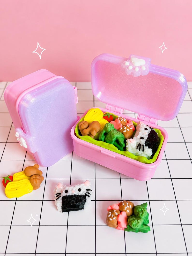 Puede incluir: Dos cajas bento de pl&aacute;stico rosa en miniatura con tapas brillantes y un cierre de huella blanca. Una caja est&aacute; abierta y muestra peque&ntilde;as gomas de borrar con forma de comida, incluyendo br&oacute;coli, bolas de arroz y carne. Hay gomas sueltas cerca.