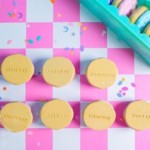 Pode incluir: Sete objetos circulares amarelos com os dias da semana gravados, dispostos sobre uma superf&iacute;cie quadriculada rosa e branca. Os dias da semana s&atilde;o segunda, ter&ccedil;a, quarta, quinta, sexta, s&aacute;bado e domingo. O fundo &eacute; decorado com confetes.