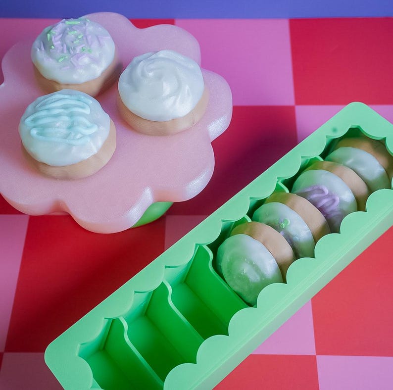 Pode incluir: Uma cole&ccedil;&atilde;o de biscoitos coloridos e gelados. Tr&ecirc;s biscoitos est&atilde;o em um suporte rosa em forma de flor, enquanto outros est&atilde;o dispostos em um recipiente verde com bordas onduladas. Os biscoitos apresentam cobertura pastel em tons de azul, branco e roxo.