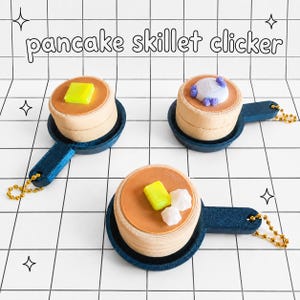 May include: Three miniature pancake skillet clickers. Each pancake has a brown top, a tan base, and sits in a dark blue skillet. One has a yellow butter square, another has a white and purple topping, and the last has a yellow butter square and white sugar cubes. The text "pancake skillet clicker" is above the pancakes.