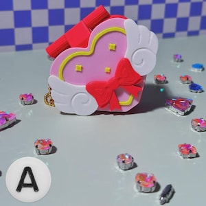 May include: A pink heart-shaped trinket box with a red bow and white wings. The box has a yellow outline and small gold squares. A red lipstick-shaped object is attached to the top. Rhinestones and a white circle with the letter "A" are scattered around.