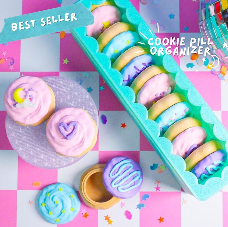 Pode incluir: Uma imagem l&uacute;dica mostra um organizador de comprimidos em forma de biscoito turquesa cheio de biscoitos em tons pastel. Biscoitos adicionais com cobertura decorativa s&atilde;o dispostos em um prato lavanda e um pequeno recipiente aberto. O texto "BEST SELLER" e "COOKIE PILL ORGANIZER" s&atilde;o vis&iacute;veis.