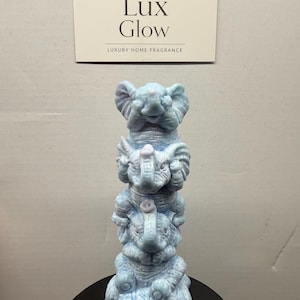 Puede incluir: Una figura de elefante azul claro apilada. La figura está hecha de tres elefantes, uno encima del otro. El producto es de Lux Glow, Luxury Home Fragrance.