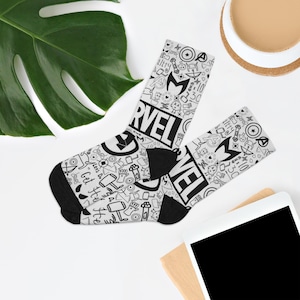 May include: A pair of white socks with a black heel and toe. The socks feature a Marvel comic book theme with the word "Marvel" in large black letters. The socks are covered in small black and white illustrations of comic book characters and symbols.