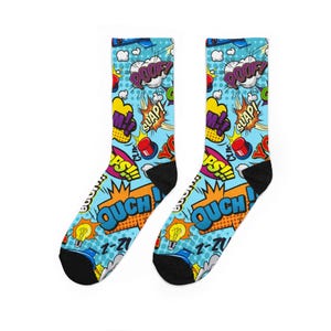 May include: A pair of crew socks with a comic book theme. The socks feature a light blue background with colorful comic book sound effects like "POOF!" and "OUCH!" along with other graphics. The socks have black heels and toes.
