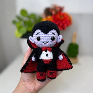 May include: A crocheted vampire doll featuring a pale lavender face, black hair, and black eyes. The doll wears a black suit with a white shirt front, a red bowtie, and a black cape with a red interior lining. It has red shoes with black cross-stitch details.