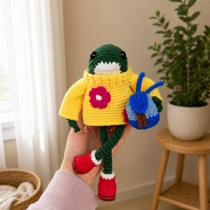 May include: A crocheted frog doll with black bead eyes, wearing a yellow sweater with a pink flower appliqué. The frog has long green legs, red boots with white trim, and carries a small blue and brown backpack with two blue antennae.