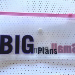 May include: A clear plastic zippered pouch with the words "BIG plans small" in black and purple letters. The zipper is pink, and the pouch has a white button closure. The background is a white surface with small red hearts.