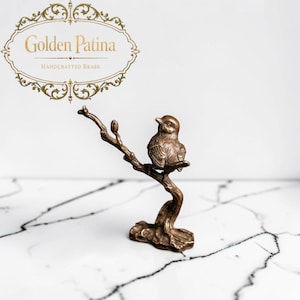 May include: A brass figurine of a small bird perched on a textured tree branch. The bird is depicted with detailed feathers and a rounded body. The branch extends upwards with a small bud. The text Golden Patina Handcrafted Brass is visible.