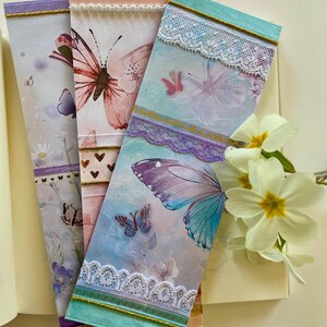 Puede incluir: Tres marcapáginas decorativos con mariposas, adornados con encaje y cintas. Los marcapáginas presentan ilustraciones de mariposas en tonos rosa, morado y azul. Cada marcapáginas tiene un diseño diferente, con ribetes dorados y detalles delicados.
