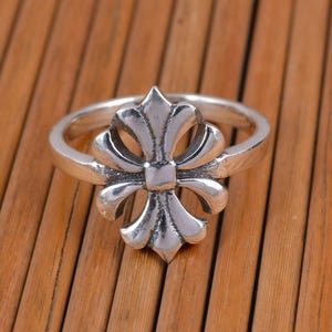 Puede incluir: Un anillo plateado con un diseño de flor de lis. El anillo presenta un patrón floral simétrico y detallado en el centro, con un acabado pulido. El anillo está sobre un fondo de madera.