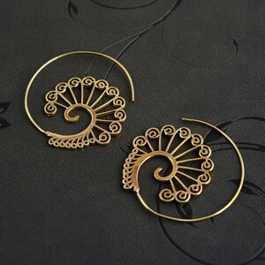 May include: A pair of gold-coloured spiral earrings. Each earring has a decorative, fan-like design with intricate details. The earrings have a circular, open-hoop design, and are displayed against a dark background.