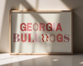 May include: A framed print with the words "GEORGIA BULLDOGS" in red and pink patterned letters. The letters are set against a cream-colored background, and the frame is a light wood color. The print is displayed on a white surface.