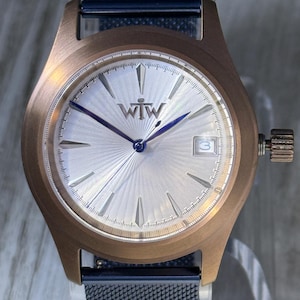 May include: A wristwatch with a bronze-colored case and a silver face. The watch has blue hands and a date display at the 3 o'clock position. The watch band is a dark blue mesh. The watch face has the letters "WtW".