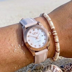 May include: A rose gold wristwatch with a light pink leather strap and a white dial featuring the letters WTW, crystal hour markers, and a date window. The watch is worn with a beaded bracelet and three gold rings on the fingers.