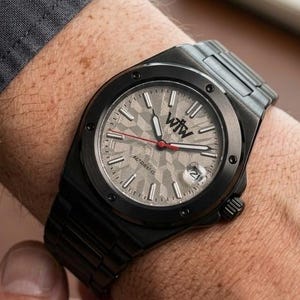 K&ouml;nnte beinhalten: Eine schwarze Armbanduhr aus Metall mit einem strukturierten hellgrauen Zifferblatt, dem Logo WtW, einem roten Sekundenzeiger und einem Datumsfenster auf der Drei-Uhr-Position. Die Uhr hat eine schwarze L&uuml;nette mit sichtbaren Schrauben und ein Gliederarmband.