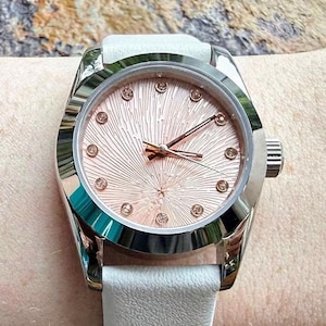 May include: A wristwatch with a polished silver-tone metal case and a white leather strap. The dial features a textured, sunburst-patterned rose gold face with small, clear crystal hour markers and slender rose gold hands for timekeeping.