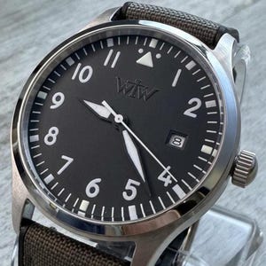 K&ouml;nnte beinhalten: Eine Armbanduhr mit rundem, silberfarbenem Metallgeh&auml;use und mattschwarzem Zifferblatt. Das Zifferblatt zeigt wei&szlig;e arabische Ziffern, ein Datumsfenster auf der Drei-Uhr-Position und den Schriftzug "WTW" unter der Zw&ouml;lf-Uhr-Markierung. Braunes Textilarmband.