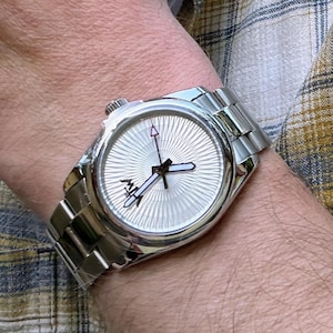 Puede incluir: Un reloj de pulsera anal&oacute;gico de tono plateado con una correa de eslabones met&aacute;licos y una esfera redonda con un patr&oacute;n de rayos de sol texturizado, manecillas de hora y minutos negras y un peque&ntilde;o detalle de logotipo negro cerca del centro.