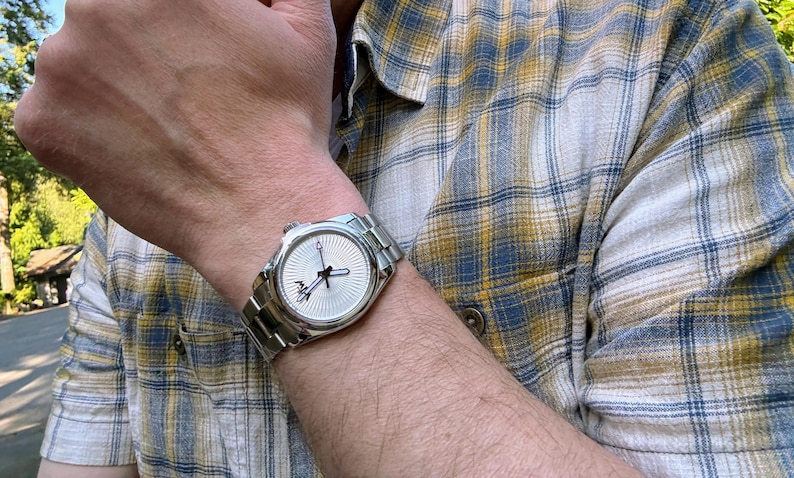 Puede incluir: Un reloj de pulsera anal&oacute;gico de tono plateado con una correa de eslabones met&aacute;licos y una esfera redonda con un patr&oacute;n de rayos de sol texturizado, manecillas de hora y minutos negras y un peque&ntilde;o detalle de logotipo negro cerca del centro.