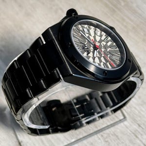 K&ouml;nnte beinhalten: Eine mattschwarze automatische Armbanduhr mit einem strukturierten grauen und schwarzen geometrischen Zifferblattmuster. Die Uhr verf&uuml;gt &uuml;ber ein schwarzes Metallgliederarmband, einen roten Sekundenzeiger, wei&szlig;e Stunden- und Minutenzeiger sowie ein Datumsfenster.