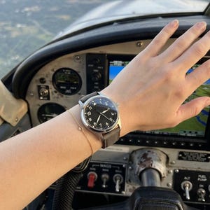 K&ouml;nnte beinhalten: Eine Armbanduhr aus Edelstahl mit schwarzem Zifferblatt, wei&szlig;en arabischen Ziffern und einem grauen Textilarmband. Die Uhr hat eine Dreiecksmarkierung auf der Zw&ouml;lf-Uhr-Position und d&uuml;nne silberne Zeiger, getragen mit einem feinen Goldkettchen.